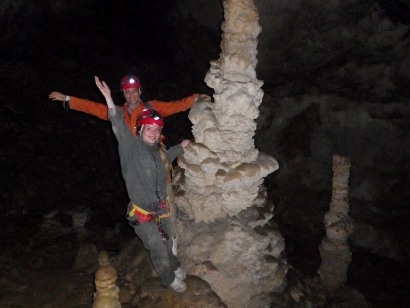 Deux spéléologues en combinaison et casque rouge posant joyeusement devant des formations calcaires dans une grotte.