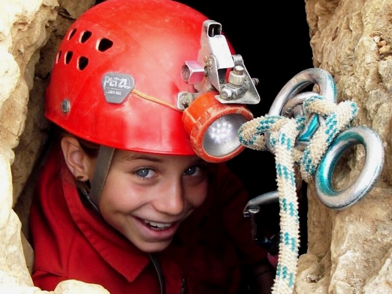 Jeune spéléologue en casque rouge avec lampe frontale, souriant dans une étroite ouverture rocheuse d'une grotte.