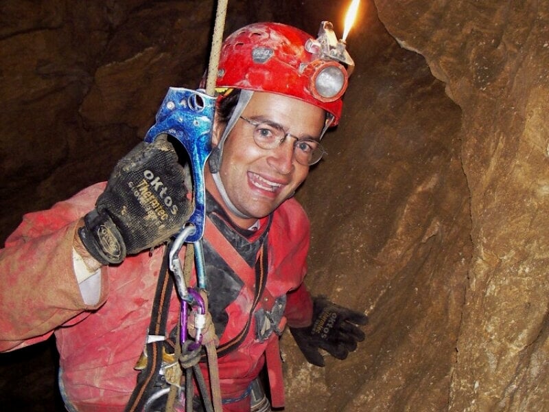 Homme en combinaison rouge et casque rouge avec lampe frontale, souriant dans une cavité rocheuse en spéléologie.