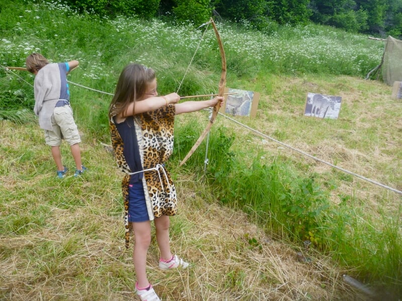 Enfant en tenue primitive lançant une lance en bois dans un champ verdoyant avec des collines en arrière-plan.