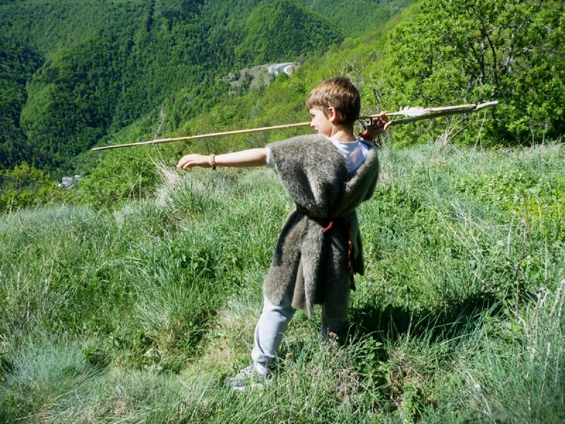 Enfant en tenue de survie tenant une longue lance, debout dans un champ verdoyant avec montagnes en arrière-plan.