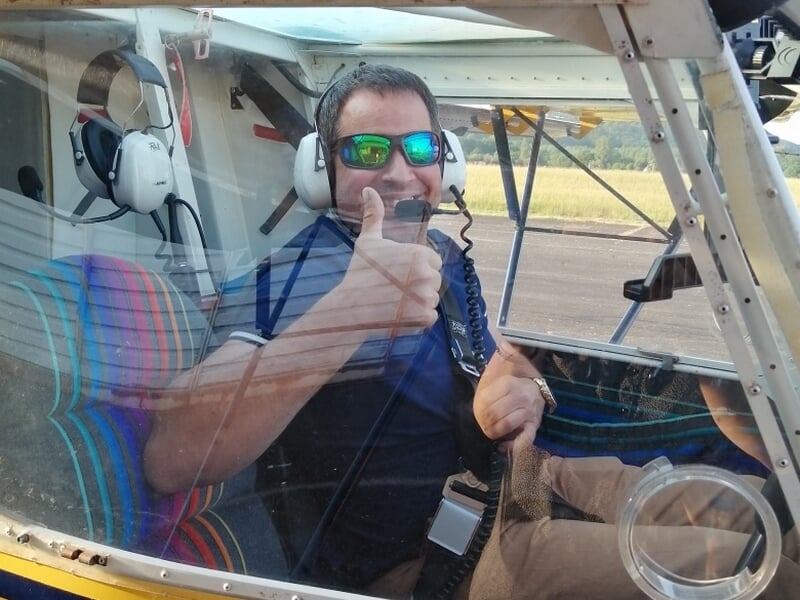 Homme souriant à l’intérieur d’un petit avion, portant casque audio et lunettes de soleil, pouce levé, vu à travers la verrière du cockpit au sol.