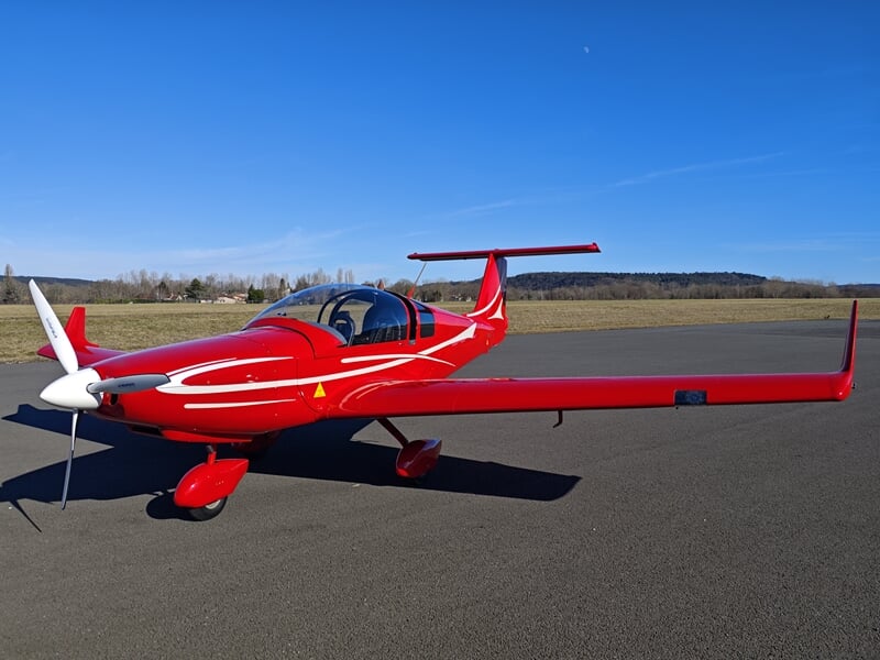 Petit avion rouge brillant avec hélice blanche, carlingue profilée et aile basse, stationné sur un tarmac sous un ciel bleu dégagé.