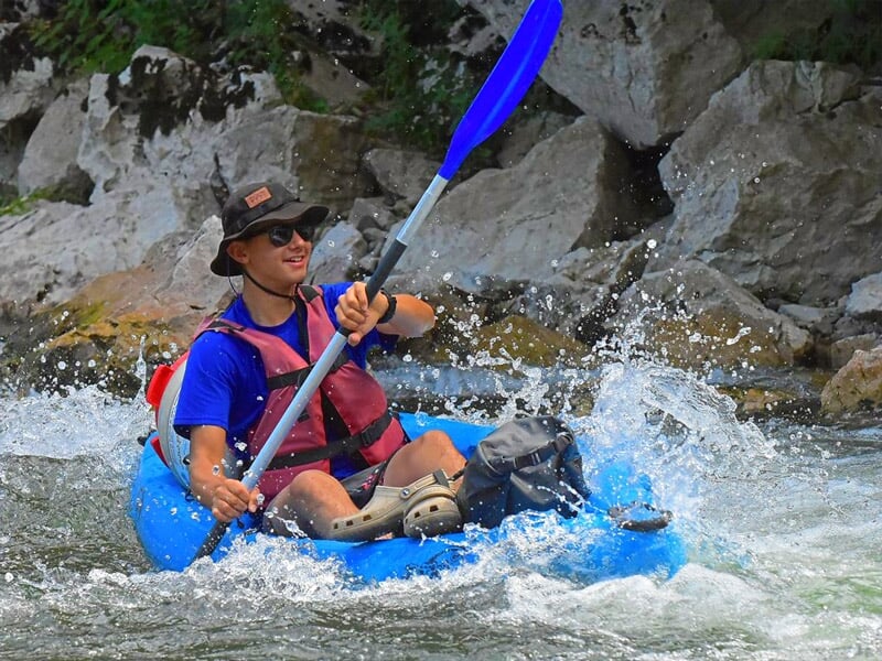 Jeune homme souriant pagayant dans un kayak bleu sur une rivière avec des rapides et des rochers.