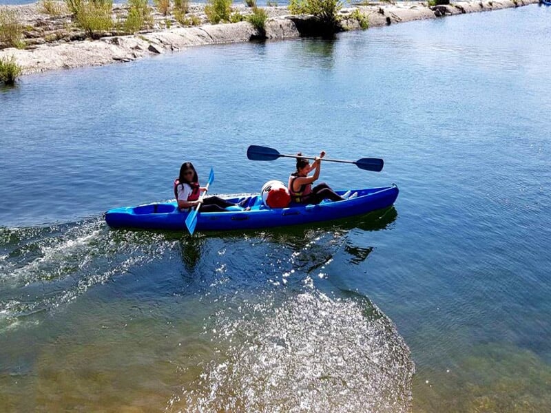 Deux enfants pagayant dans un kayak bleu sur une rivière sous un ciel bleu avec quelques nuages.