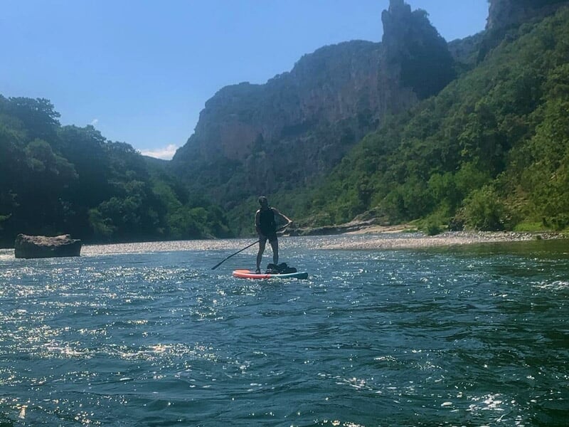Personne debout sur une planche de stand-up paddle pagayant sur une rivière ensoleillée.