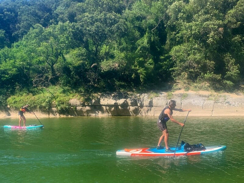 Personne pagayant sur une planche de stand-up paddle sur une rivière calme avec falaises en arrière.