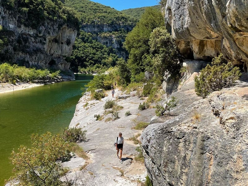 Personne marchant sur un sentier rocheux au bord d'une rivière dans un paysage naturel verdoyant.