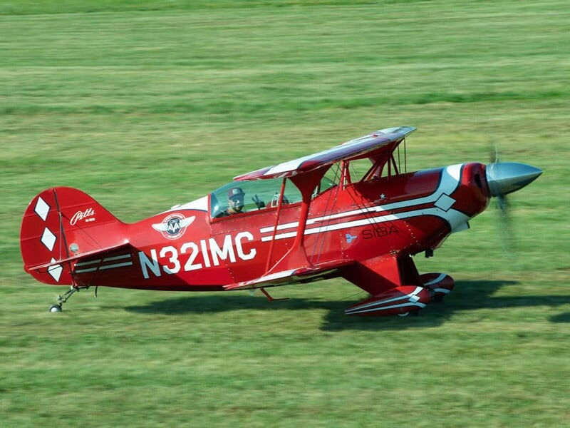 Avion de voltige rouge et blanc roulant sur une piste d'herbe verte à l'aérodrome.