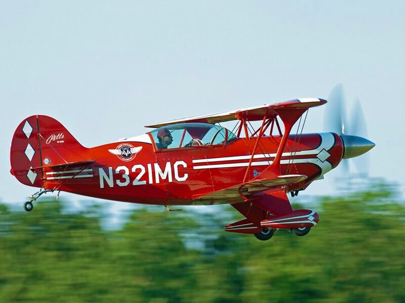 Avion de voltige rouge et blanc en vol avec un fond de verdure floue en arrière-plan.