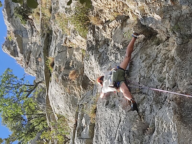 Un grimpeur en short et casque orange escalade une falaise rocheuse avec des arbres autour sous un ciel bleu.