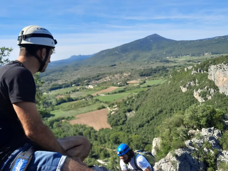 Un homme en casque blanc et t-shirt noir observe le paysage vallonné et boisé depuis un point d'escalade en hauteur.