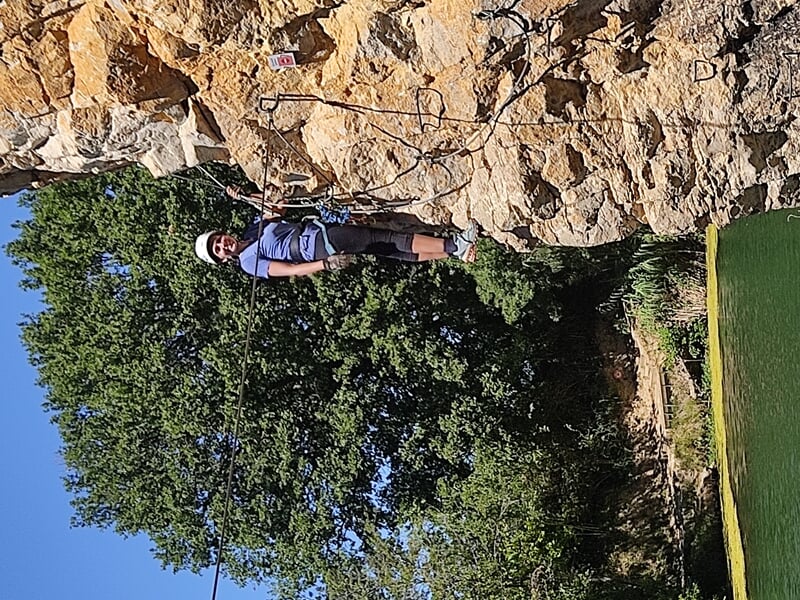 Une personne en casque blanc et équipement de via ferrata traverse un pont de câbles suspendu au-dessus d'une rivière.