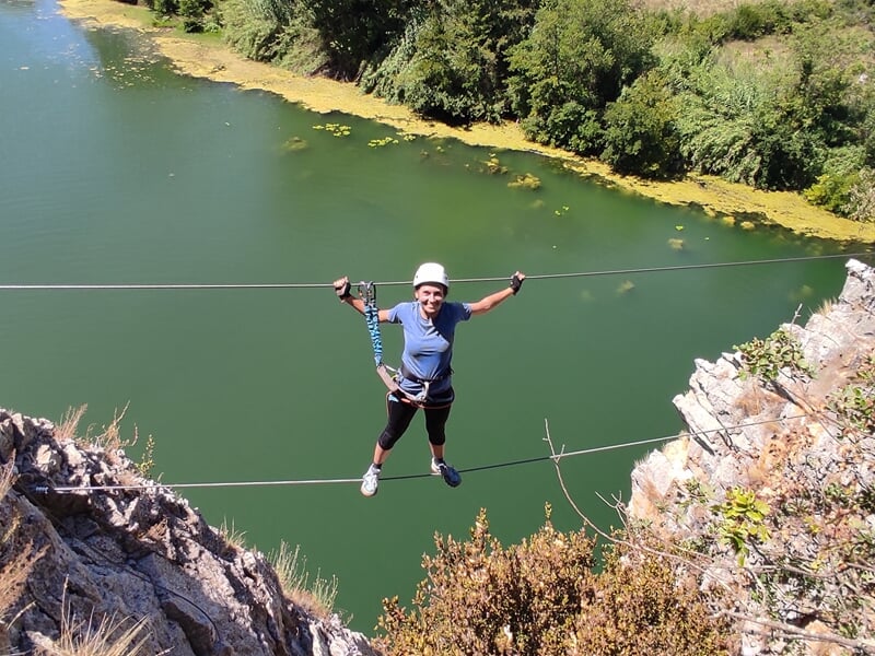Un grimpeur en casque blanc avance sur une via ferrata avec des câbles et des échelons métalliques au-dessus de l'eau.