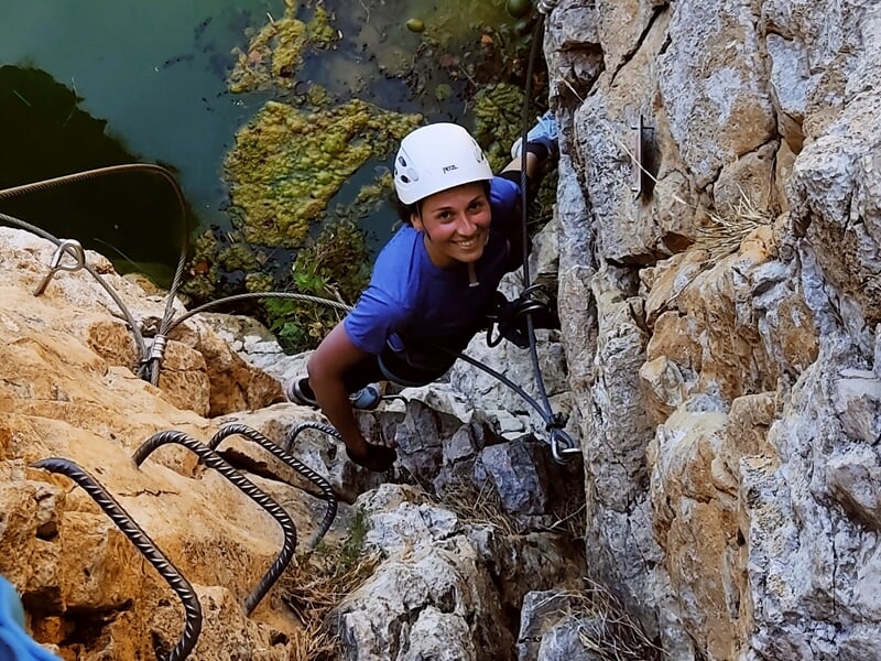 Une grimpeuse en casque blanc souriante escalade une paroi rocheuse équipée d'une via ferrata au bord d'un lac.