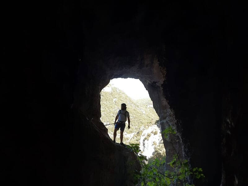 Un grimpeur en casque blanc progresse sur une via ferrata avec vue sur une rivière et des falaises rocheuses environnantes.