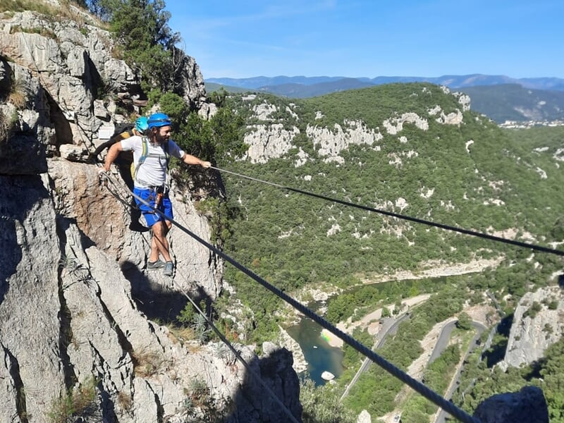 Un grimpeur en casque blanc traverse un pont de câbles suspendu au-dessus d'une rivière dans un paysage naturel.
