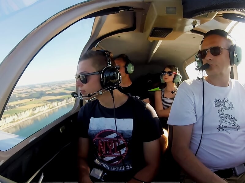 Trois personnes portant un casque audio dans un petit avion en vol au-dessus d'un paysage côtier ensoleillé.