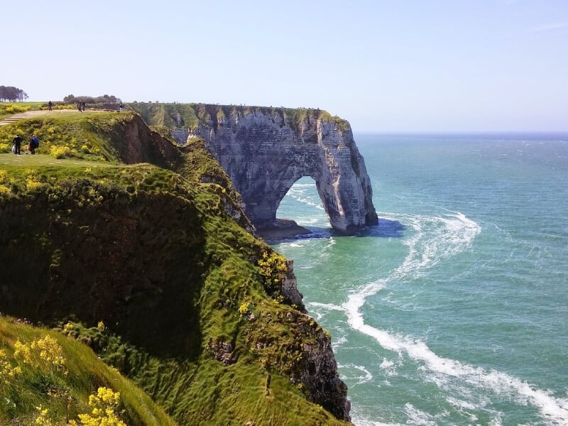 Vue panoramique d'une falaise avec une arche naturelle au bord de la mer sous un ciel bleu clair.