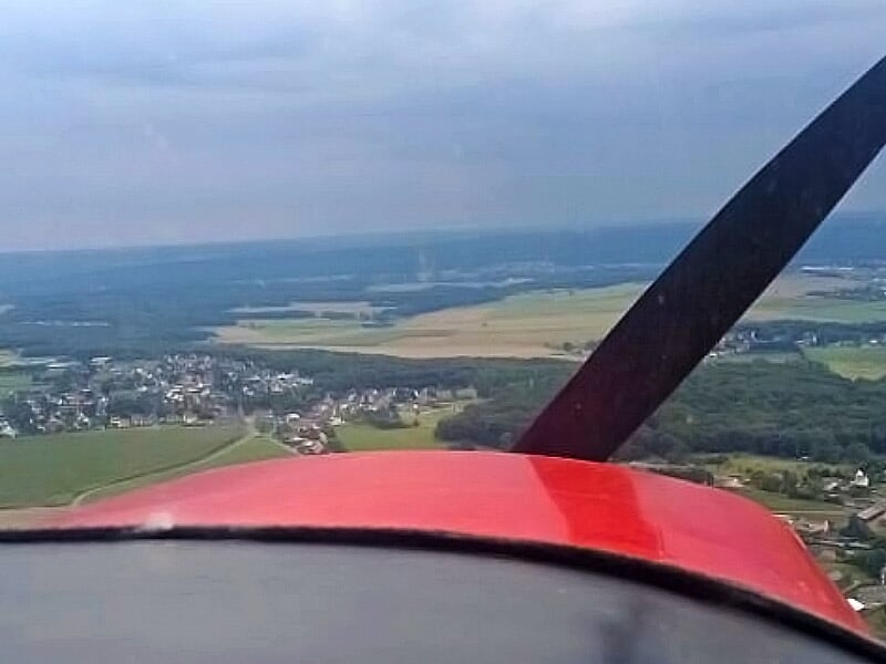 Vue depuis le cockpit d'un petit avion rouge en vol, survolant des champs et un village sous un ciel nuageux.