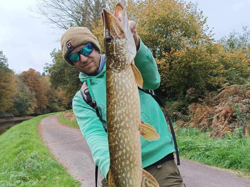 Un homme en tenue verte et lunettes de soleil tient un grand brochet capturé lors d'une sortie de pêche en nature.