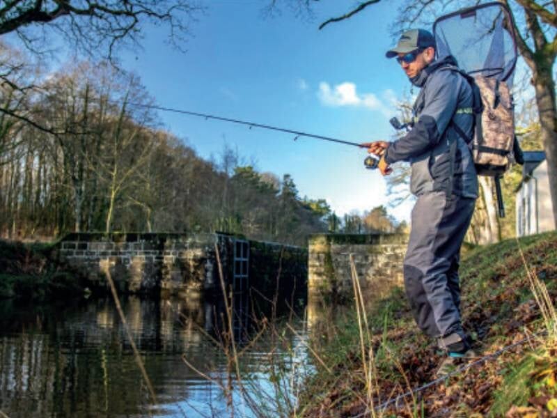 Lors d'un stage de street-fishing près de Loudéac, un pêcheur en tenue grise avec sac à dos lance sa ligne dans une rivière calme près d'une écluse entourée d'arbres en automne. 