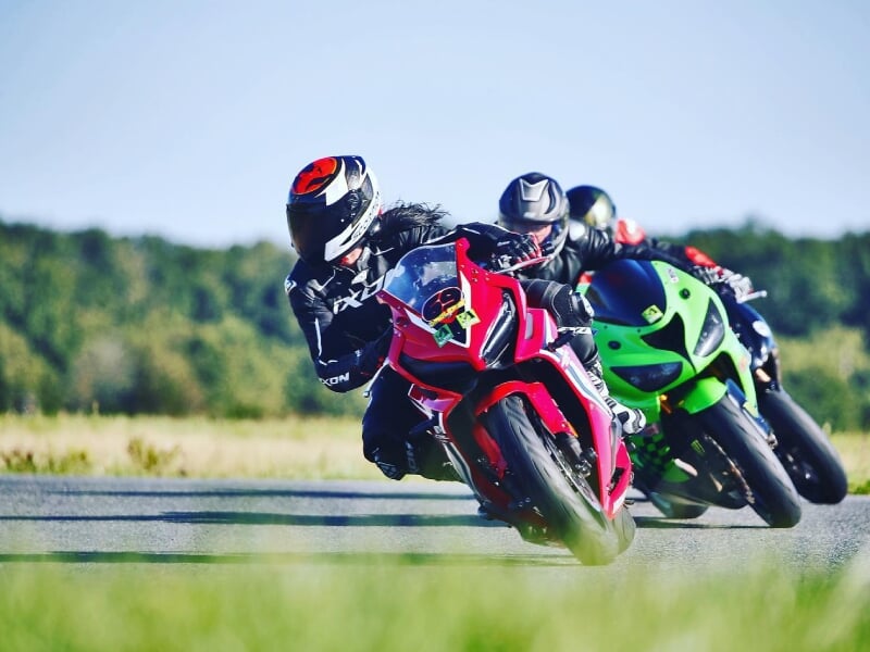 Trois motards en course, prenant un virage sur une piste de circuit, conduisant des motos rouges et vertes, avec des arbres et de l'herbe en arrière-plan