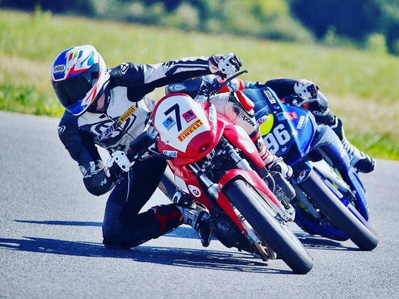 Deux motards en course, prenant un virage serré sur un circuit asphalté. Le pilote en tête porte une combinaison noire et blanche, et conduit une moto rouge et blanche avec le numéro 7