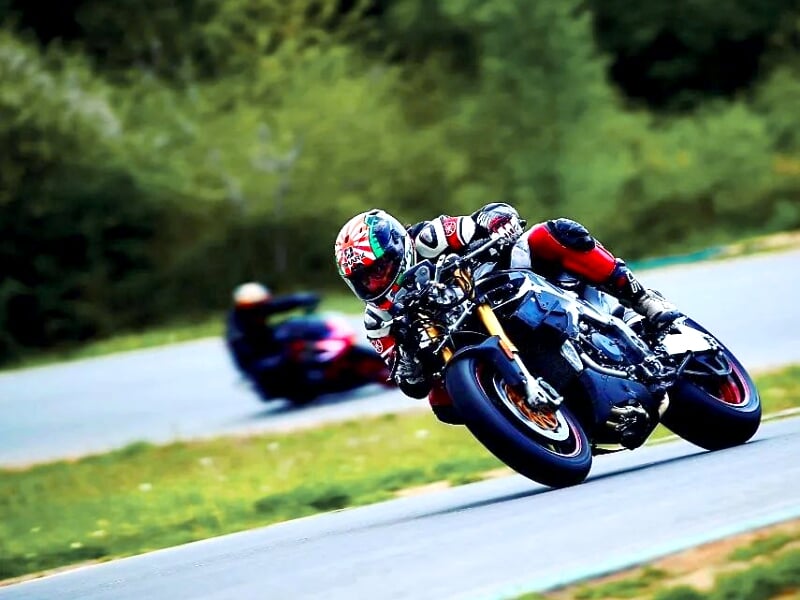 Motard en combinaison noire et rouge, inclinant fortement sa moto dans un virage sur circuit, suivi de près par un autre pilote en combinaison bleue