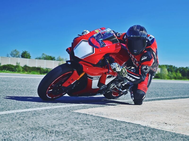 Motard en combinaison rouge et noire, prenant un virage serré sur une moto de course rouge sur un circuit, fond de ciel bleu et végétation