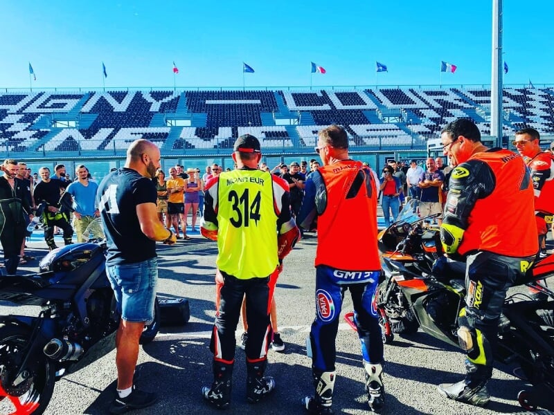 Instructeurs et participants d'un stage de moto sur circuit, portant des chasubles jaunes et orange numérotées, se tenant en groupe sur la piste devant des gradins remplis