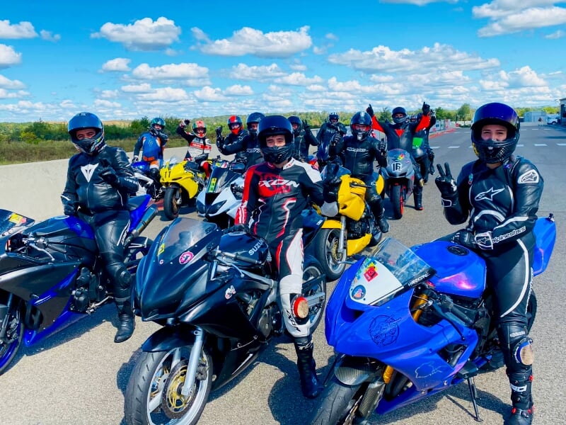 Groupe de motards en combinaison, sur des motos sportives de différentes couleurs, posant sur une route sous un ciel bleu avec quelques nuages, certains faisant des signes de la main