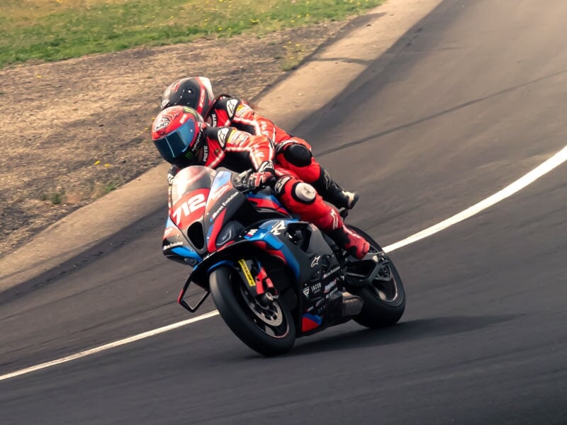 Moto de course bleue et rouge avec deux motards en tenue de course rouge, prenant un virage incliné sur une piste d'asphalte noir