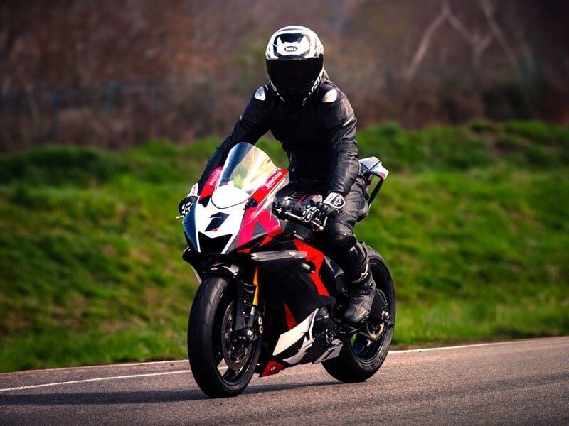 Motard en combinaison noire sur une moto rouge et blanche, roulant sur une piste avec un paysage verdoyant en arrière-plan
