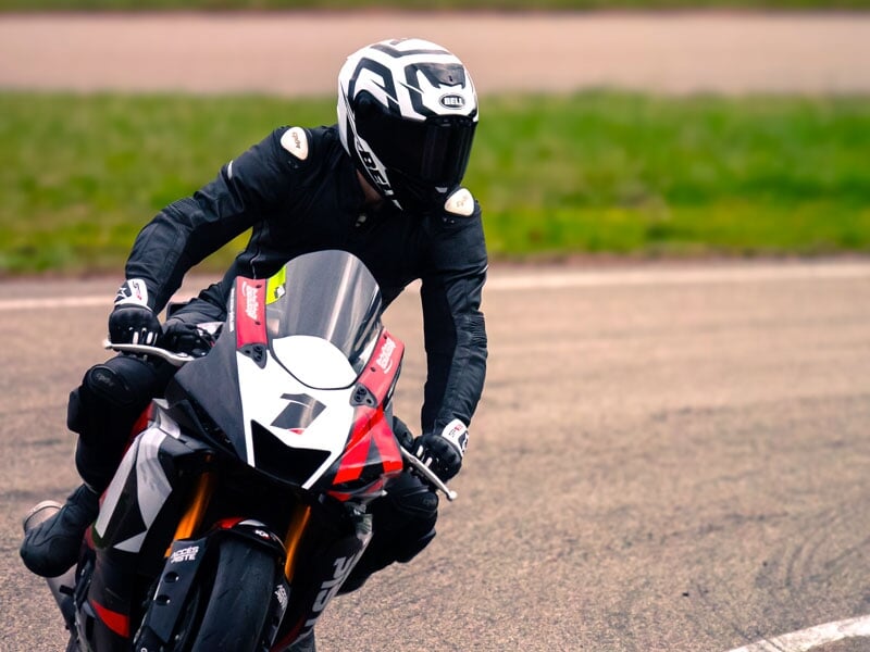 Motard en combinaison noire et casque intégral, conduisant une moto sportive rouge et blanche, prenant un virage sur une piste avec fond herbeux