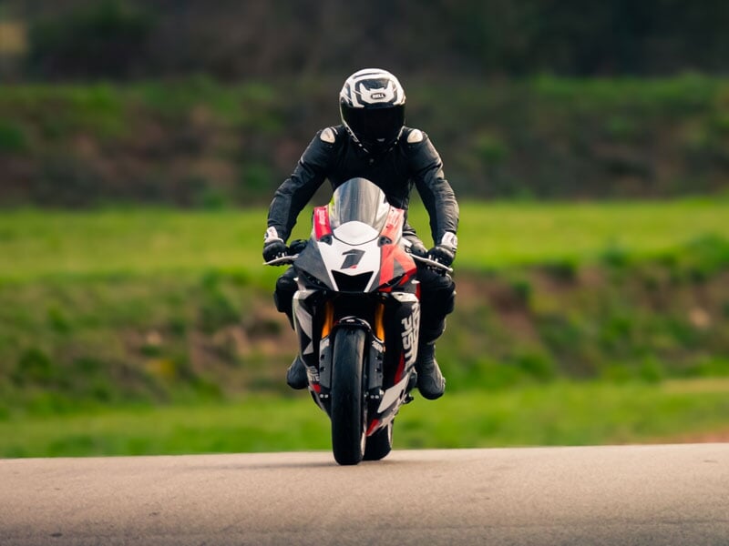 Moto sportive rouge et blanche en pleine course, vue de face, avec un motard en tenue noire sur une piste entourée de verdure