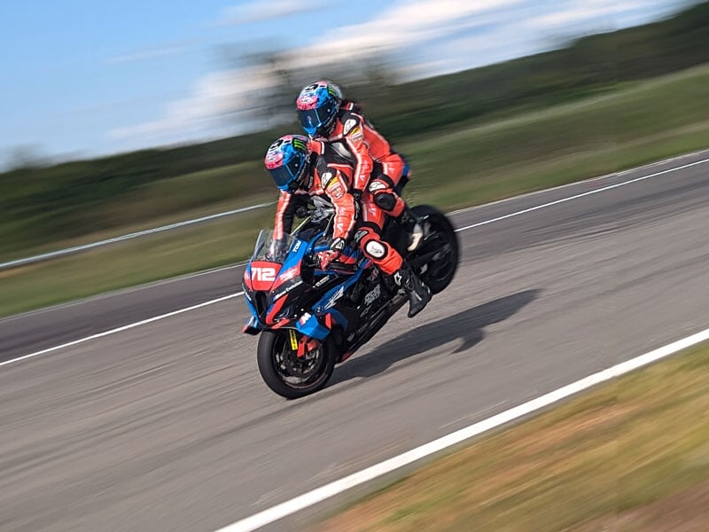 Duo de motards en combinaison rouge, exécutant un wheeling sur une moto de course bleue et rouge sur une piste, avec un léger flou de mouvement
