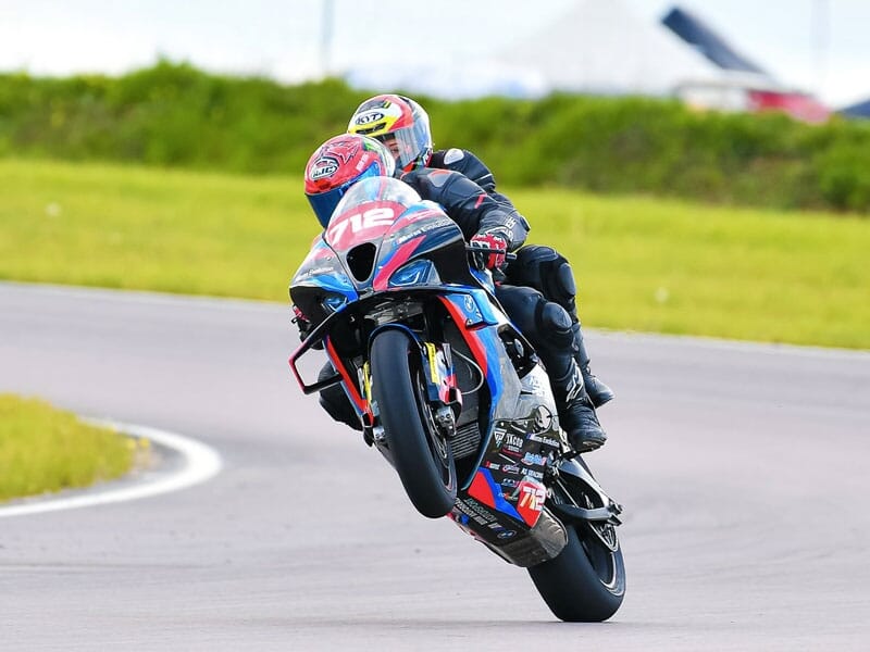 Deux motards en tenue de course sur une moto bleue et rouge, soulevant la roue avant dans un virage serré sur circuit, fond herbeux