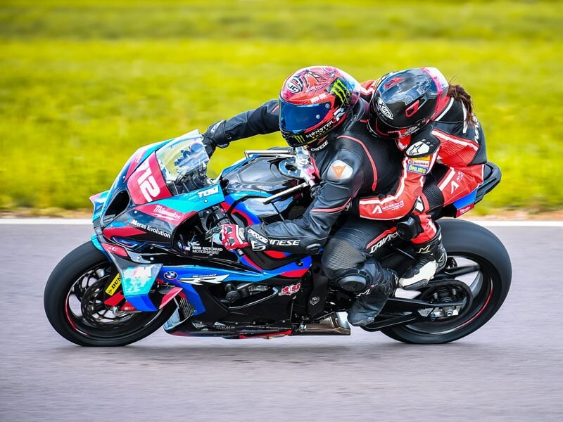 Deux motards en combinaison de course serrée, se penchant dans un virage avec une moto de course bleue et rouge, sur une piste entourée de verdure