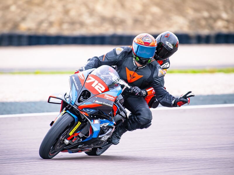 Motard et passager en combinaison noire, prenant un virage sur une moto de course bleu et rouge, le conducteur faisant un signe de paix