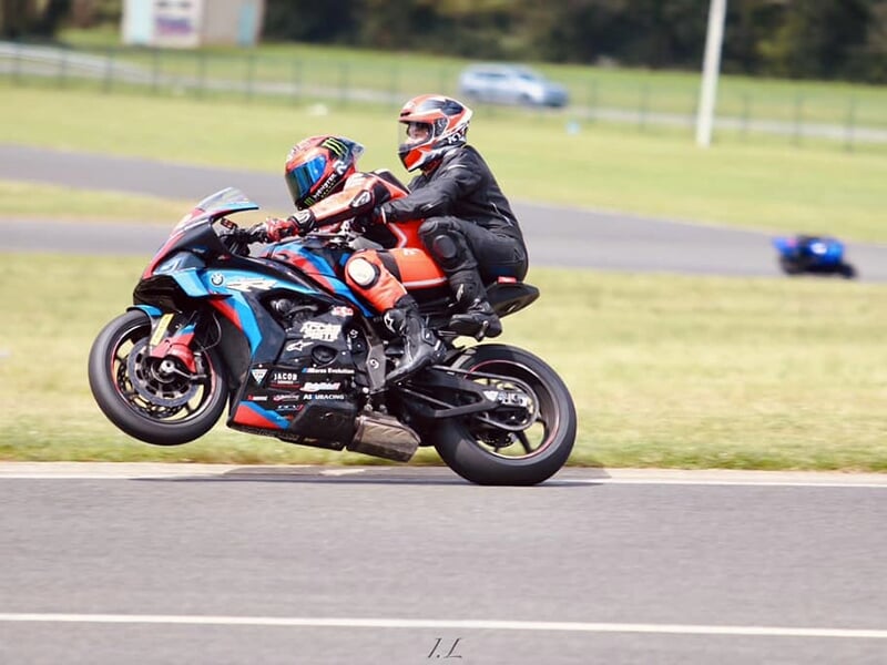 Moto de course effectuant un wheeling avec deux pilotes, tous deux vêtus de combinaisons noires et rouges, sur une piste dégagée.