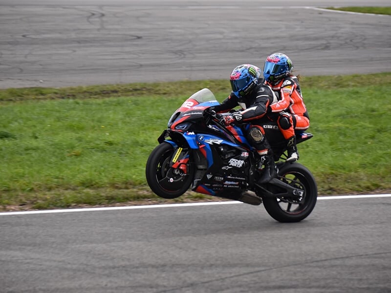 Moto numéro 712 avec deux pilotes exécutant une figure en ligne droite, le pilote arrière bien visible, dans un cadre herbeux et de piste.