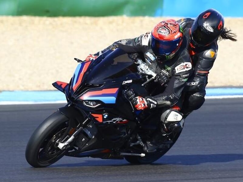 Moto en pleine action sur un circuit avec deux pilotes penchés, le pilote arrière portant une longue chevelure visible sous son casque.