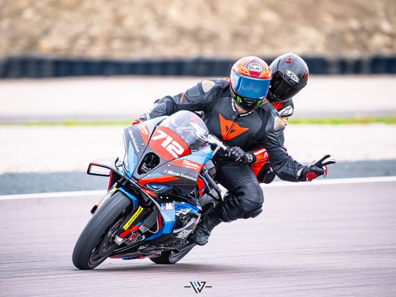 Motard en combinaison noire et passager en rouge prennent un virage serré sur une moto 712, le pilote fait un signe de paix avec les doigts.