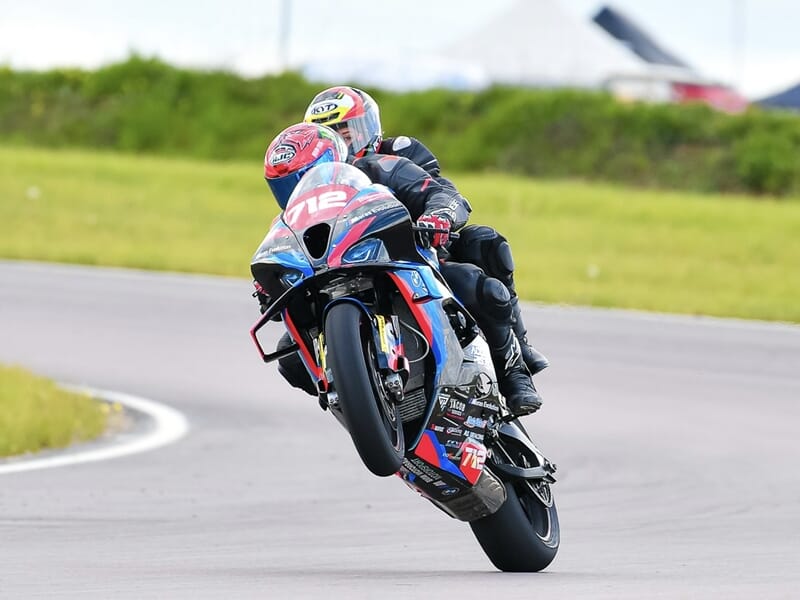 Deux motards en noir soulèvent la roue avant d’une moto de course 712 bleu et rouge dans un virage sur piste dégagée.