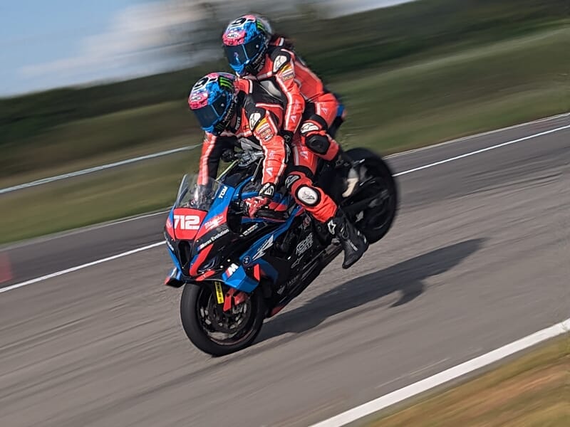 Deux motards en combinaison rouge et casque coloré effectuent une roue arrière sur une moto de course numéro 712 sur circuit asphalté flou.