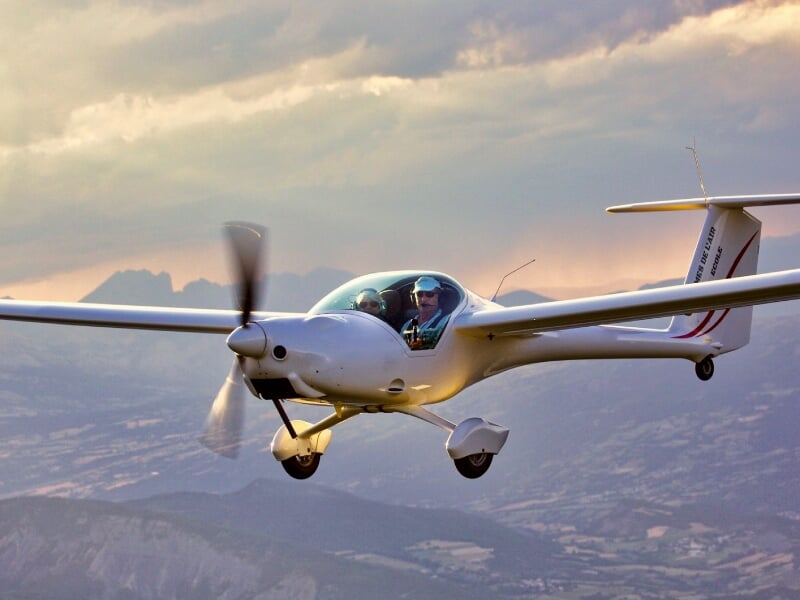 Planeur blanc en vol au-dessus des montagnes avec un pilote visible dans le cockpit sous un ciel nuageux.