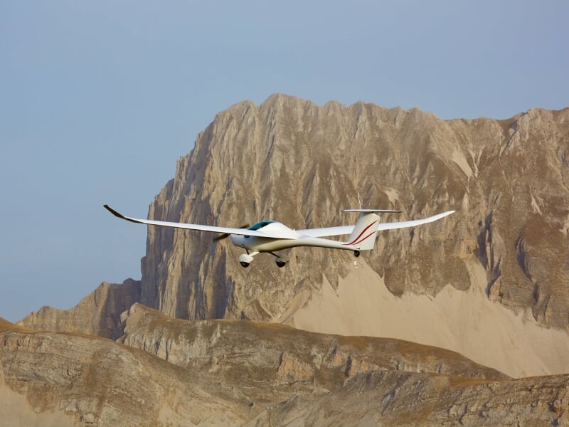 Planeur blanc survolant un paysage montagneux rocheux avec un ciel bleu clair en arrière-plan.