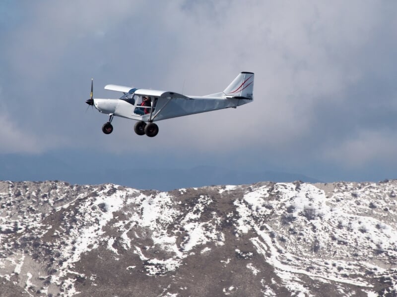 Avion ULM blanc en vol au-dessus d'un terrain montagneux avec des sommets enneigés sous un ciel bleu.