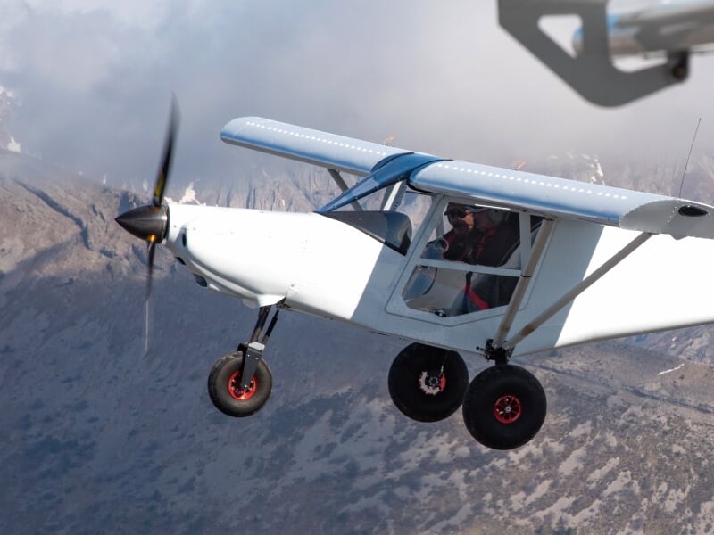 Avion ULM blanc en vol avec un pilote visible, survolant un paysage montagneux rocheux sous un ciel clair.